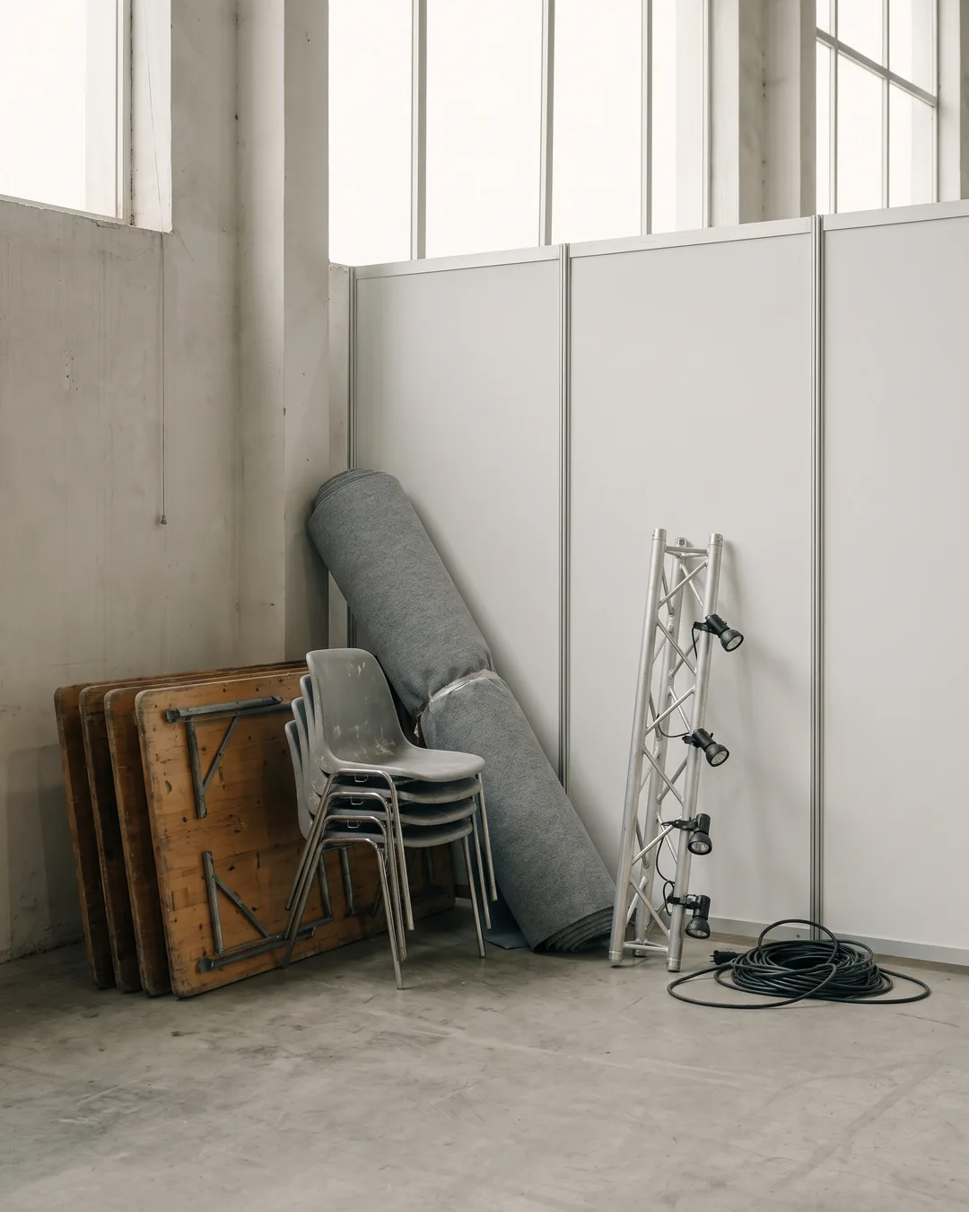 Folding tables, chairs, a roll of carpet, a rail of spotlights and a power cable, set in front of the panels of a booth.